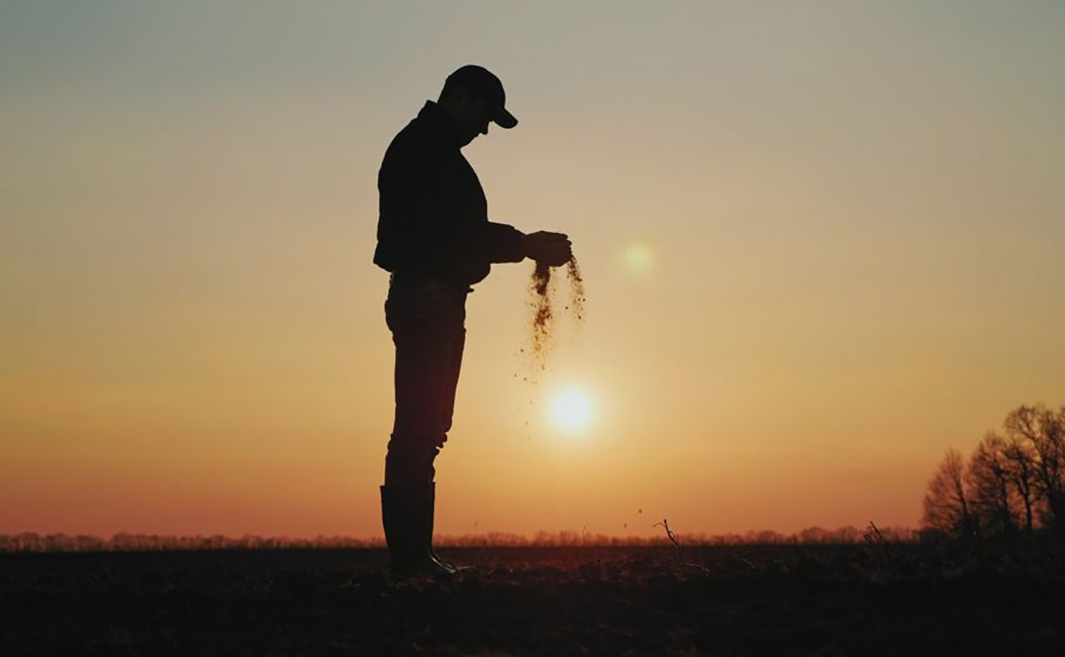 Silhouette of farmer standing in a field at sunset, holding soil in hands.
